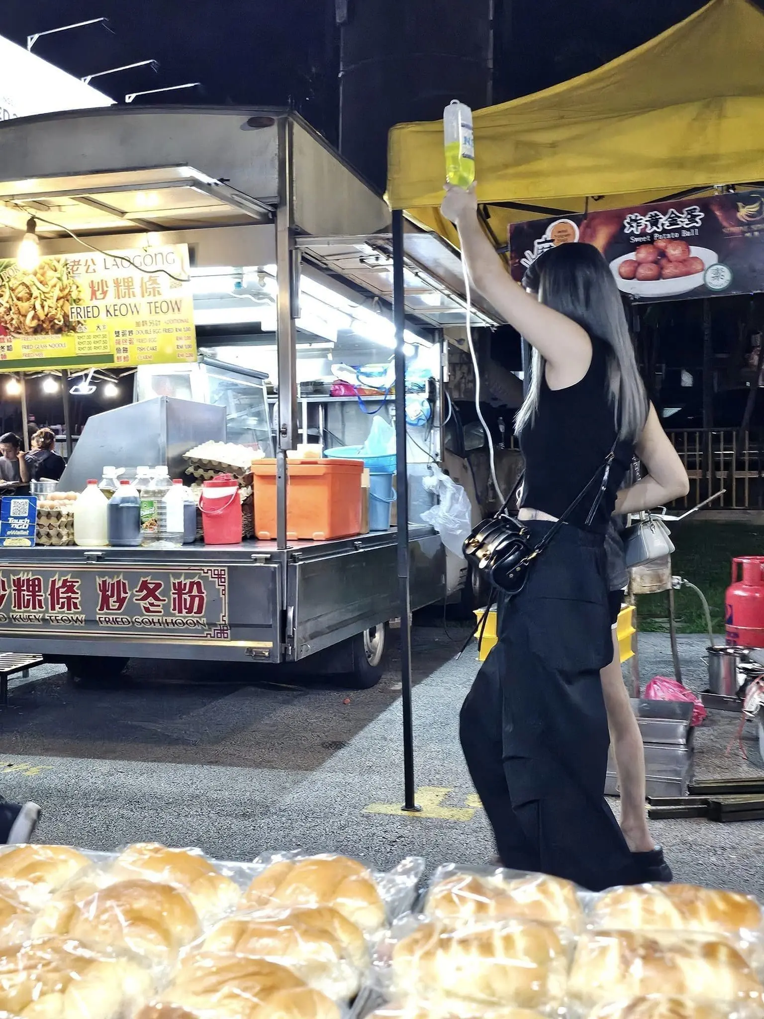 Woman's friend helps her lift IV bag while visiting Taman Connaught night market