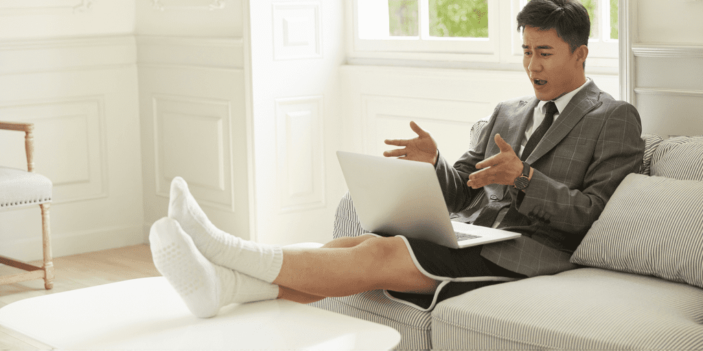 Man in a pair of shorts sitting on a sofa looking confused with a laptop in front of him.