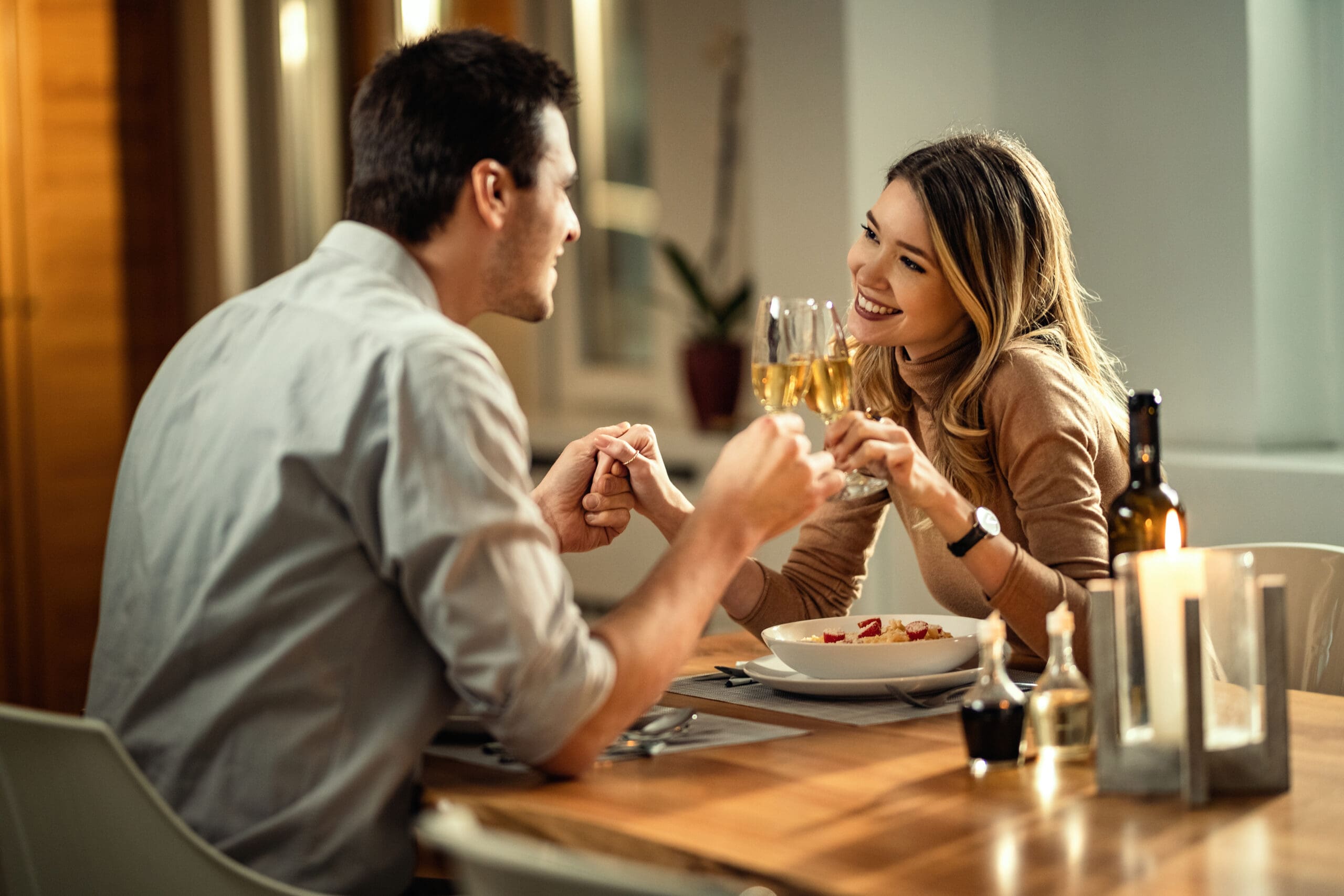 Happy woman and her boyfriend holding hands while toasting with Champagne while having dinner at dining table.