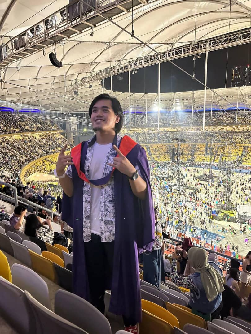 Msian graduate wearing his robe at a Coldplay concert, taking a picture before the concert starts